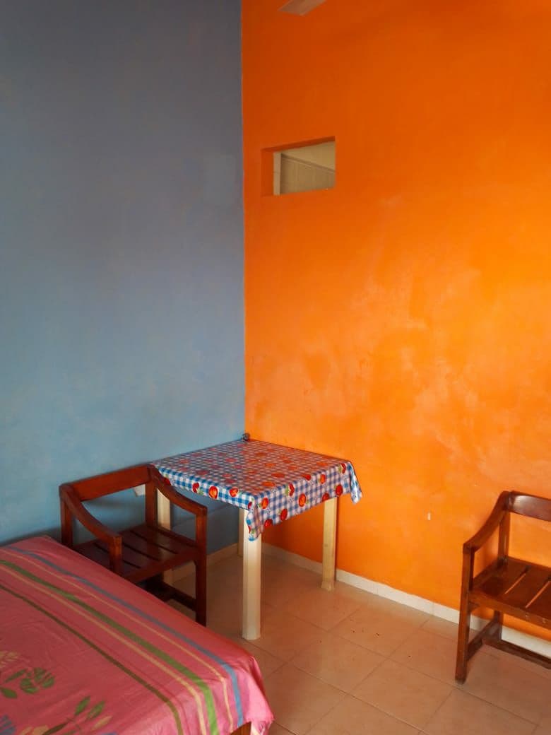 A room features a vibrant orange accent wall paired with a calming blue wall. A small table with a checkered tablecloth is positioned between two wooden chairs. A single bed with a colorful striped blanket is situated against one wall, offering a cozy space.
