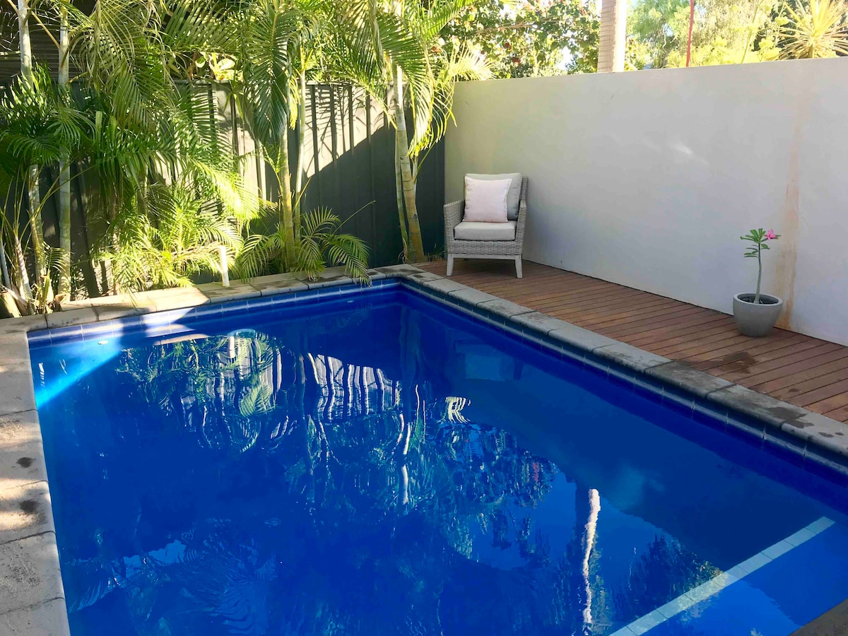 A private pool is surrounded by lush greenery, creating a serene environment. A wooden deck is positioned beside the pool, featuring a comfortable chair and a small potted plant, enhancing the tranquil setting.