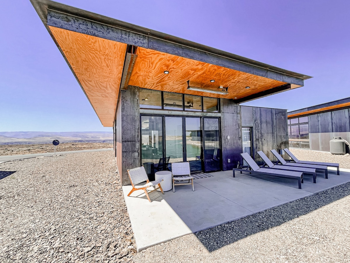 A modern vacation house is presented with a distinctive metal exterior, featuring a spacious outdoor patio. Lounge chairs are arranged along the concrete surface, with a seating area positioned beneath overhanging wood panels. Vast views of the surrounding landscape are visible through large glass doors.