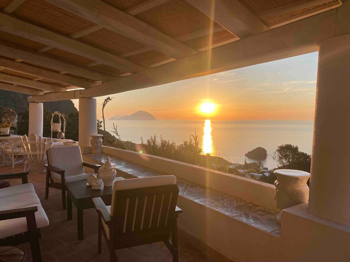 A spacious covered veranda features comfortable seating facing the sea. The warm glow of the sunset reflects on the water, with the silhouettes of islands visible in the distance. Elegant pottery adds a decorative touch along the edge of the veranda.