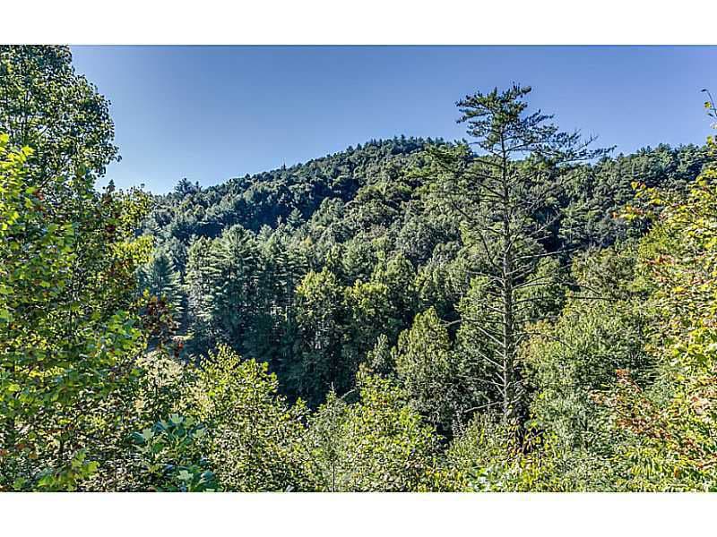A lush landscape of dense trees features varying shades of green, highlighting the natural beauty of the surrounding mountains. The clear blue sky serves as a backdrop, creating a serene atmosphere that emphasizes the peacefulness of the retreat.