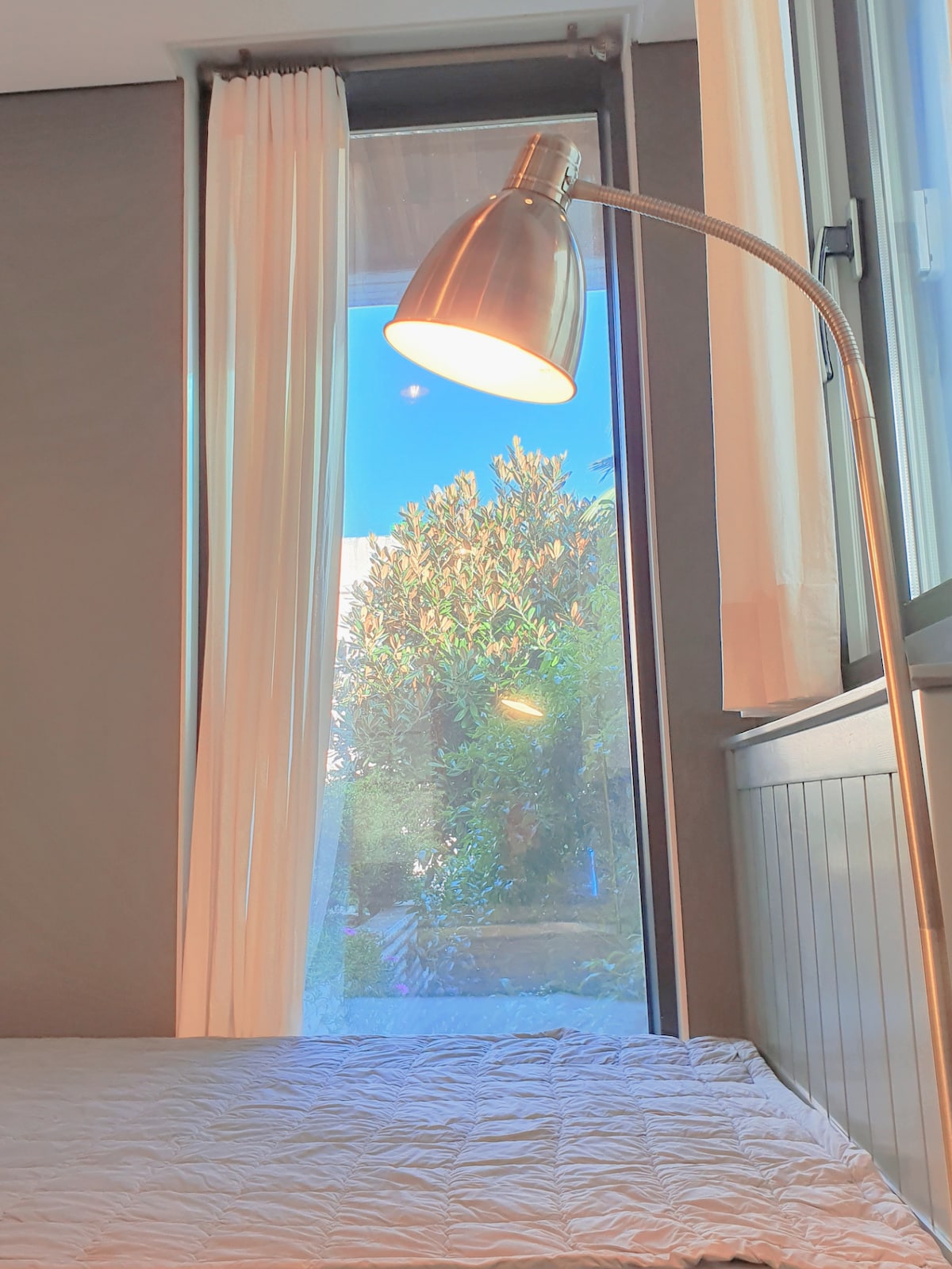 A floor lamp stands next to a bed, illuminating the soft bedding. A large window reveals a view of greenery outside, with sunlight casting a warm glow through sheer curtains. Natural light brightens the room, enhancing the serene atmosphere.