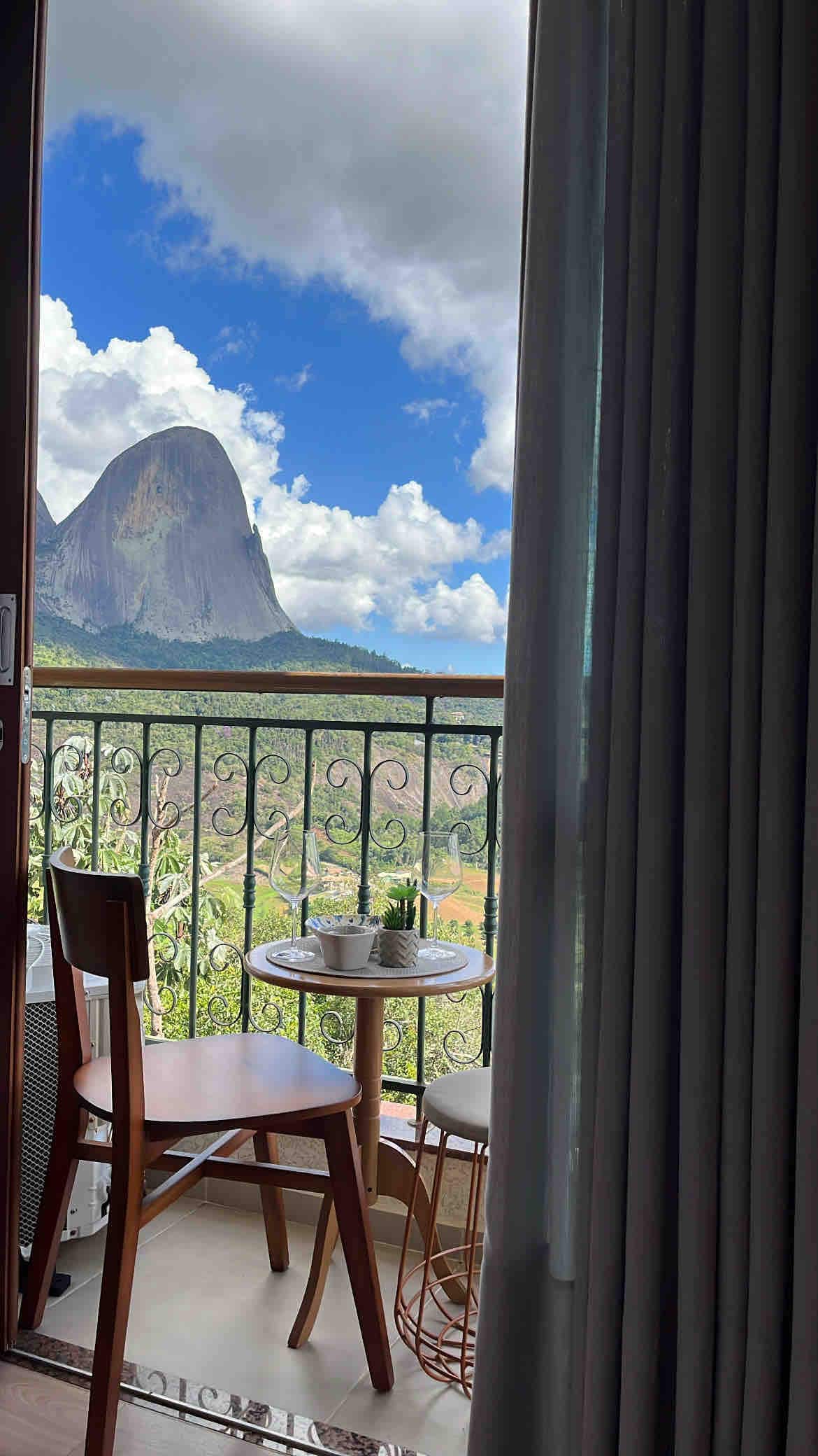 A private balcony offers a view of the iconic Pedra Azul mountain. A small round table with two chairs is positioned on the balcony, inviting relaxation while enjoying the scenery. Lush greenery surrounds the area, complementing the serene atmosphere.