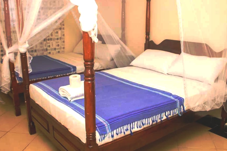 The sleeping area features two spacious beds, each draped with a white mosquito net. The beds are accompanied by fresh linens and a blue throw blanket. A rolled towel is neatly placed on one bed, enhancing the tidy appearance of the room.