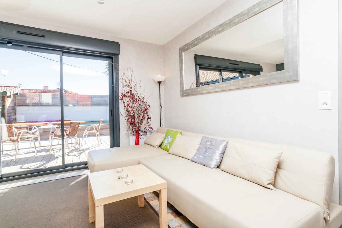 A light-filled living area features a modern white sectional sofa adorned with decorative cushions. A small wooden table sits on a patterned rug. Large glass doors provide access to an outdoor patio, while a mirror enhances the sense of space.