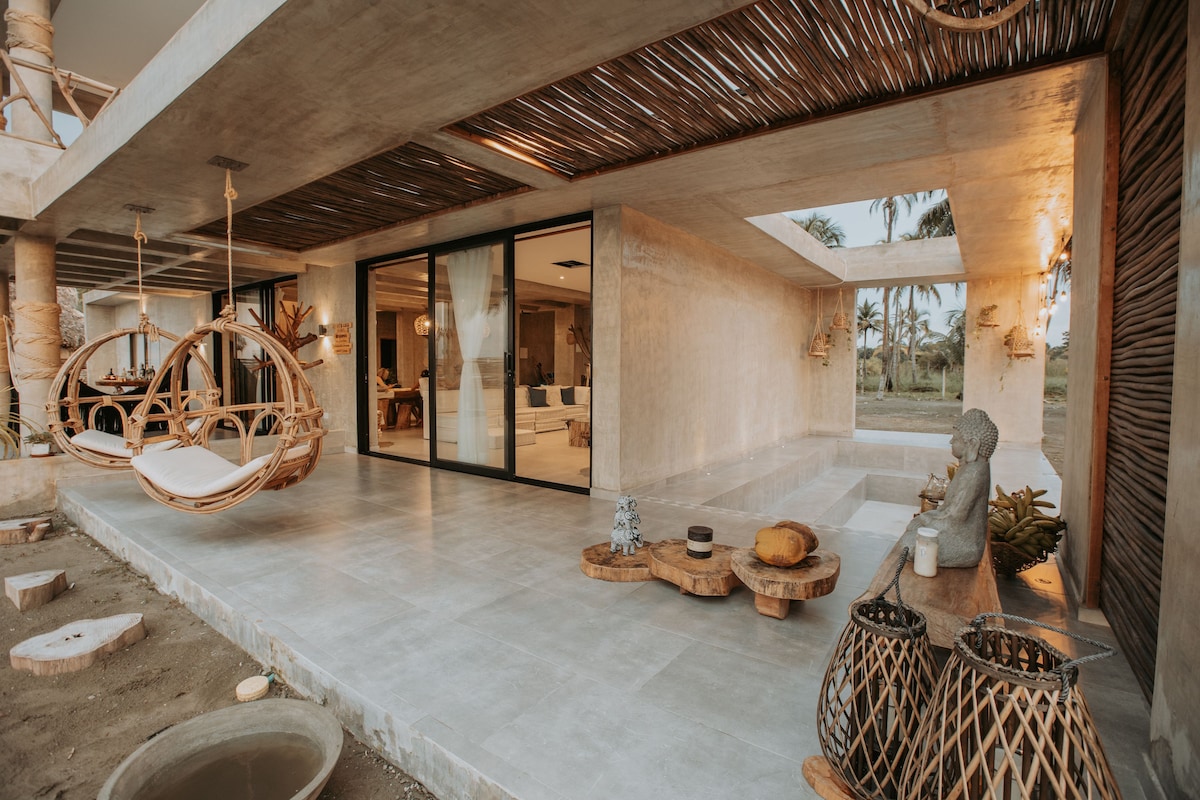 A spacious outdoor area features a modern design with large glass doors opening to the interior. Two hanging chairs provide comfortable seating, while natural materials are used throughout the space, including wooden accents and decorative baskets. Surrounding greenery enhances the tranquil atmosphere.