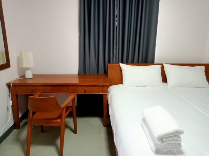 The room features a spacious bed with crisp linens and two white towels neatly arranged on top. A wooden desk with a matching chair is positioned next to the bed, while a dresser mirror reflects the soft light from a nearby lamp. Floor-to-ceiling curtains provide privacy.