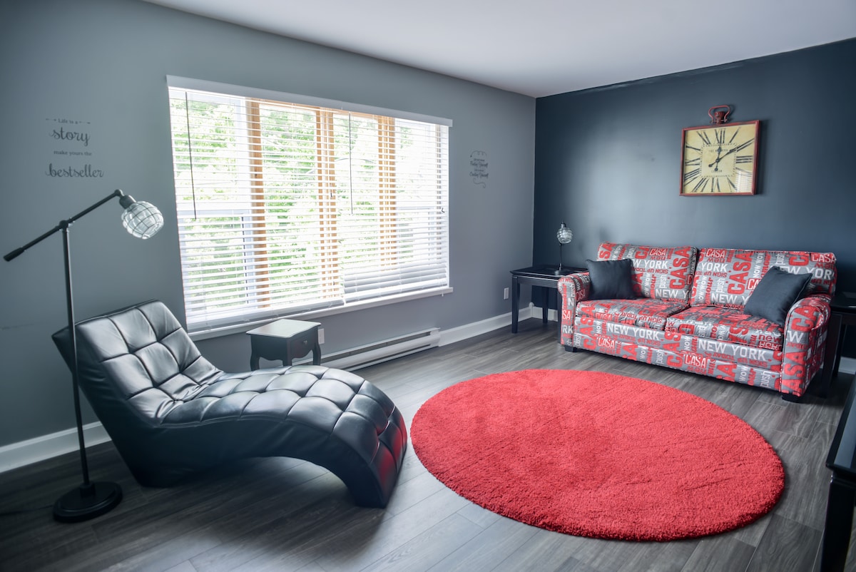 A contemporary lounge area features a black chaise lounge and a vibrant red circular rug on a wood floor. Large windows with blinds allow natural light to brighten the space. A sofa, decorated in a New York theme, complements a modern black side table with a lamp.