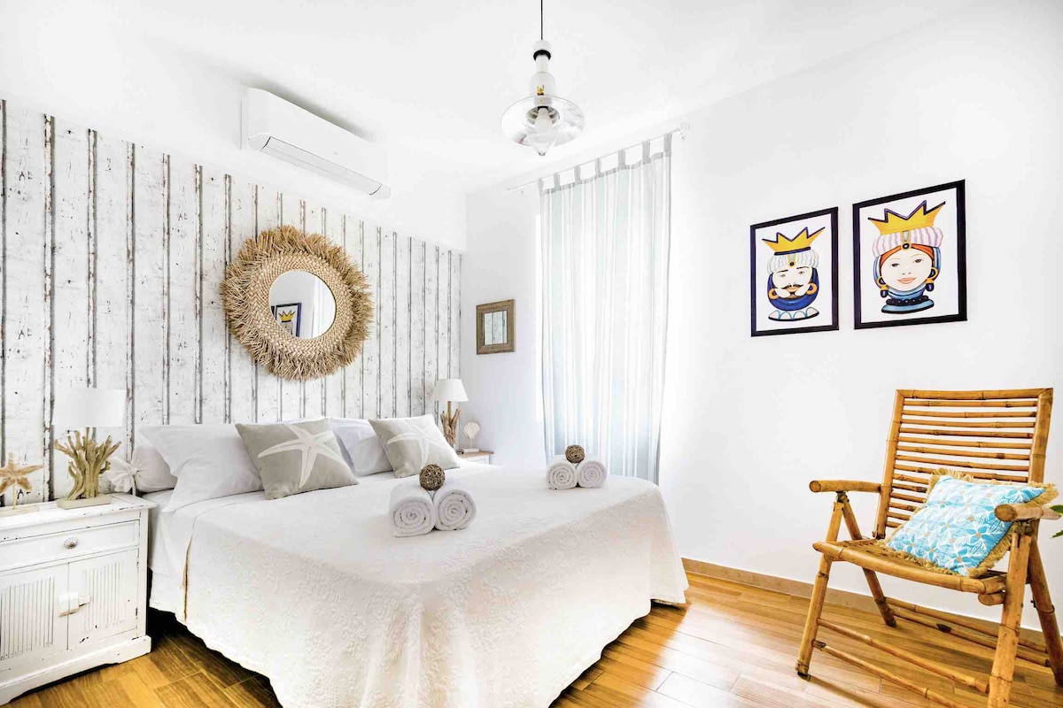 A bright bedroom features light-colored wooden panels and a spacious bed adorned with neutral linens. Decorative items, including a round mirror and wall art, enhance the decor. A bamboo chair is positioned near a window, and bath towels are neatly arranged on the bed.