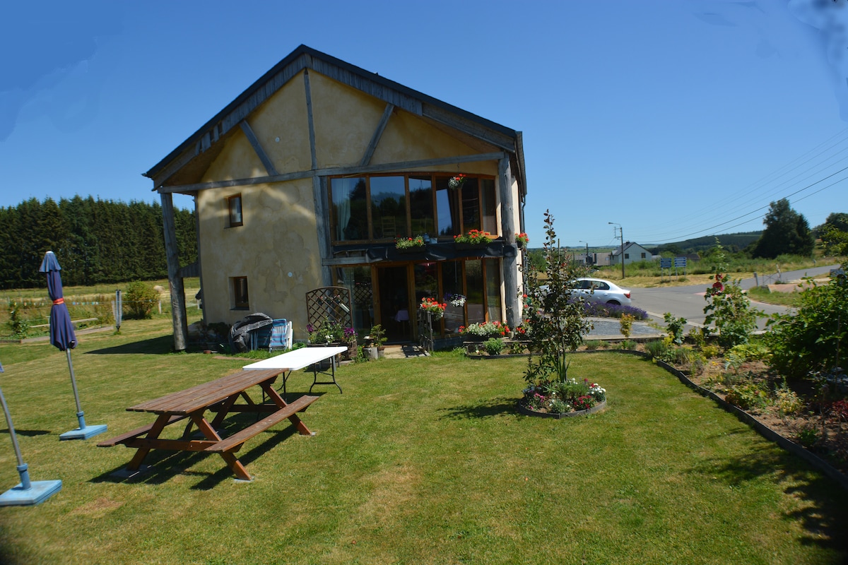 The exterior of a two-story house is presented, featuring a rustic design with wooden accents. A well-kept garden surrounds the property, showcasing green grass and blooming flowers. Picnic tables and umbrellas are visible on the lawn, providing an inviting spot for outdoor relaxation.