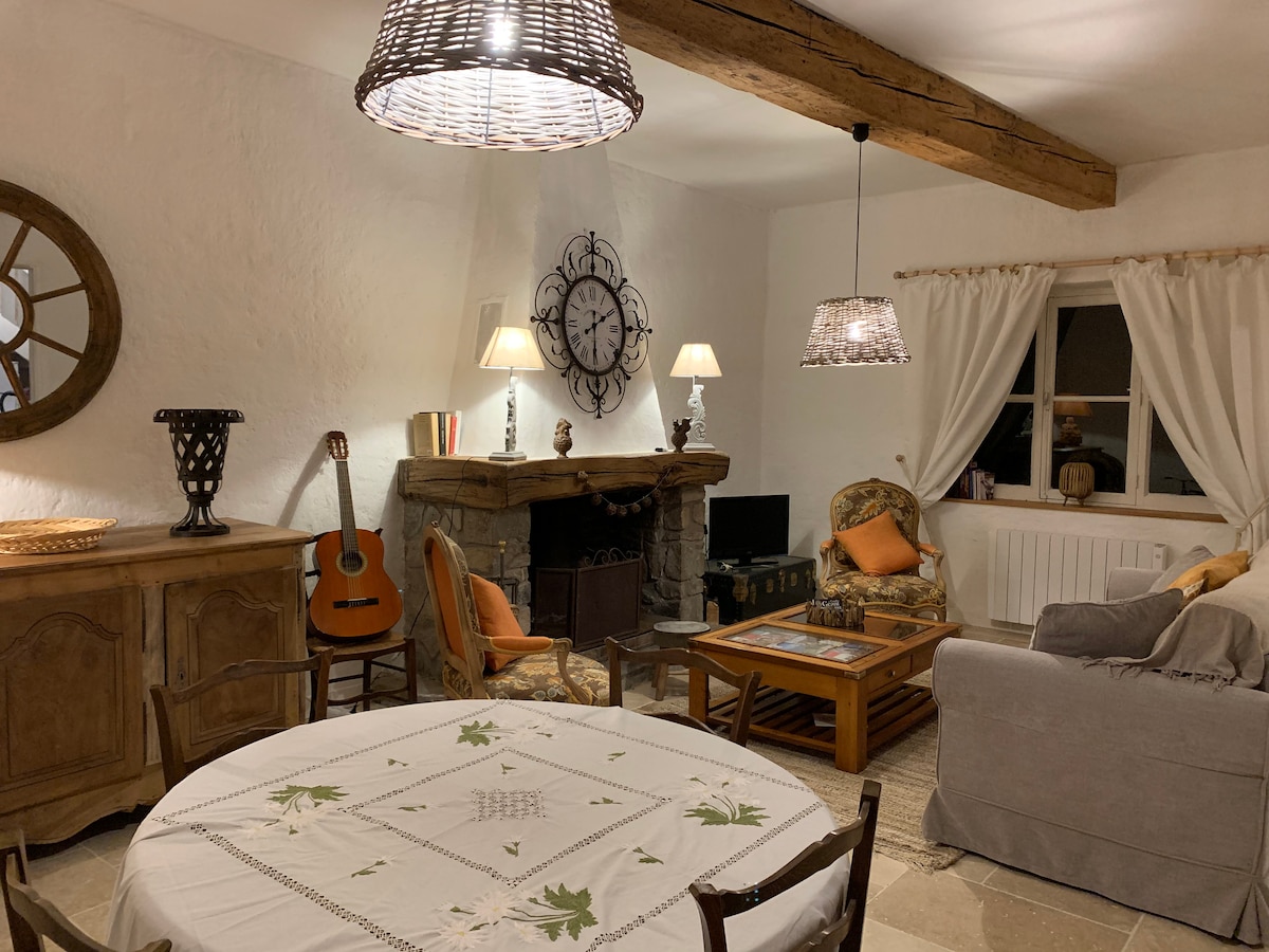 A cozy living room features a wooden beam ceiling and various seating options, including a plush gray sofa and two accent chairs. A round dining table is set with a floral tablecloth. A large clock hangs above a stone fireplace, complemented by soft lighting from woven pendant lights.