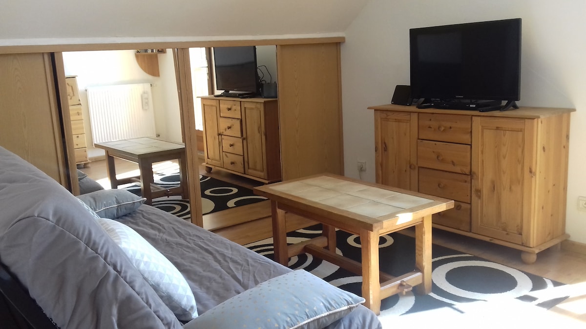 A living area is presented with wooden furniture, including a coffee table and a media console with a television. A sofa with light blue pillows is visible, and a patterned area rug covers the wooden floor beneath. Natural light is streaming in from a nearby window.