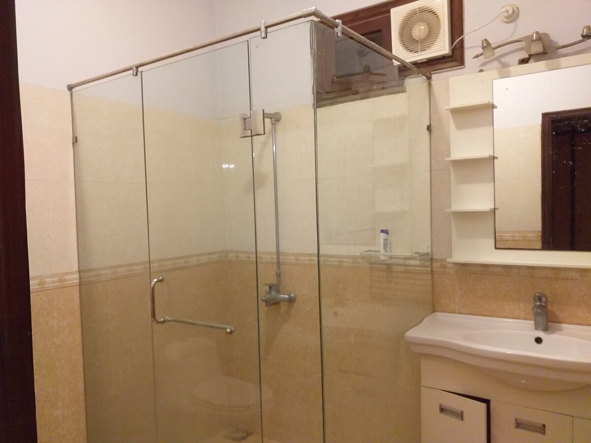 A spacious bathroom features a glass-enclosed shower with a silver showerhead. Light-colored tiles adorn the walls, and a modern vanity with a sink is present. A mirror is mounted above the sink, and storage shelves are positioned nearby, offering a tidy space.