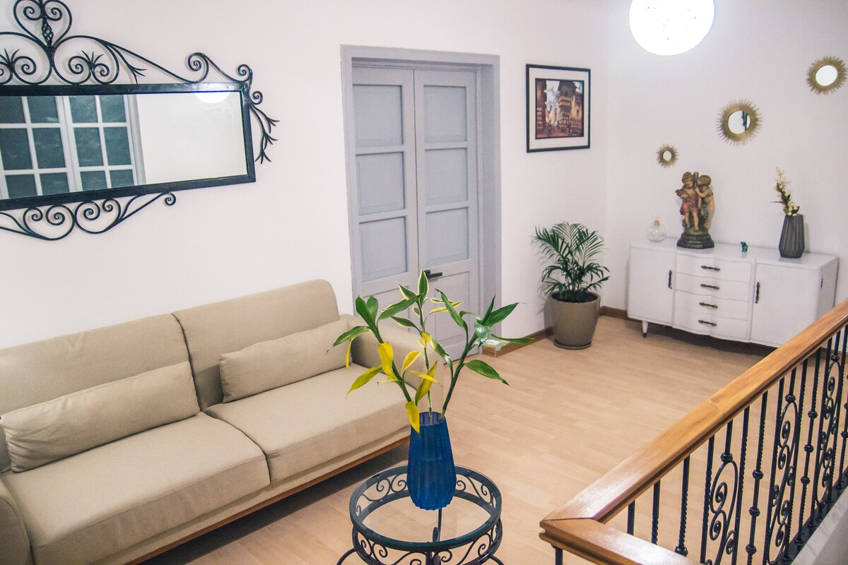 A bright living area is highlighted by a soft beige sofa and a decorative side table with a vase of yellow flowers. A mirror adorns one wall, while artwork and small sculptures add visual interest. Natural light fills the space, creating a welcoming ambiance.