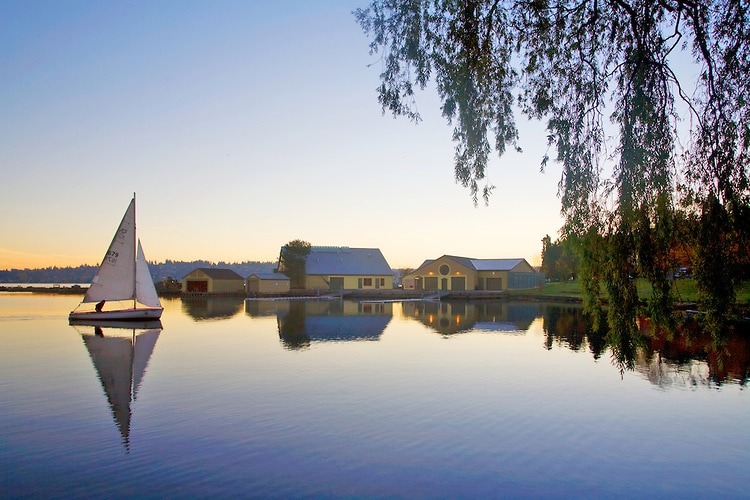 A tranquil lakeside scene captures a white sailboat gliding over calm waters, with reflections of the surrounding trees and buildings. The soft light of dawn illuminates the sky, creating a serene atmosphere, while a gentle silhouette is formed by the shoreline's structures.