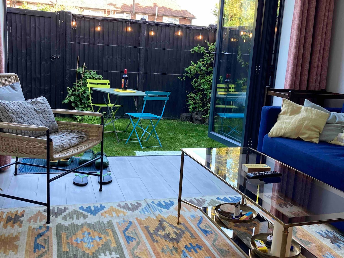 A bright living area features a modern blue sofa and a glass coffee table, with a colorful rug below. A large glass door opens to a garden with green grass and two outdoor chairs at a small table. The space is well-lit by natural light.