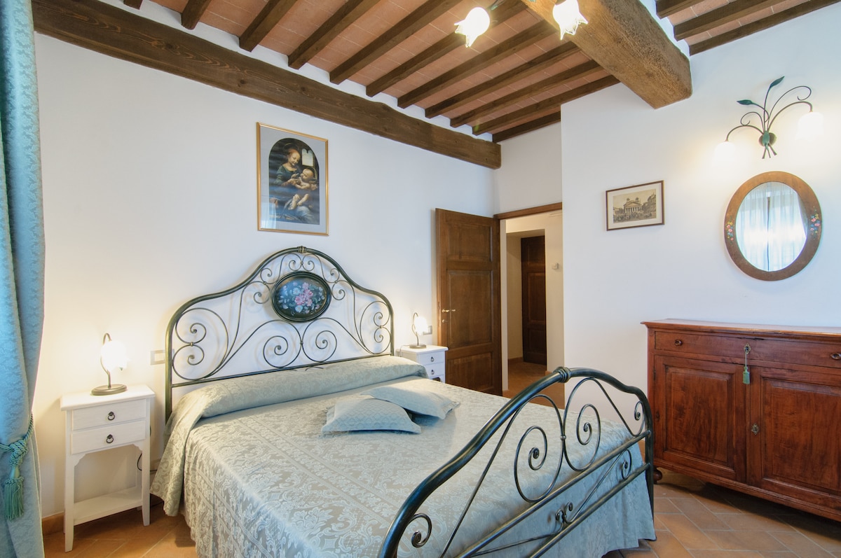 A spacious bedroom features a wrought iron bed with a light blue decorative cover. Traditional wooden beams run across the ceiling, while soft lighting is provided by bedside lamps. A wooden cabinet stands against the wall, accompanied by a small side table and a framed artwork.