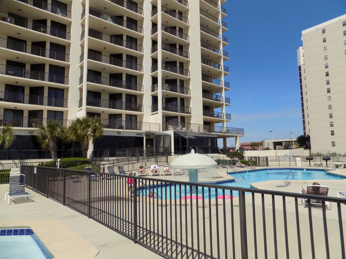 The image captures a well-maintained outdoor pool area surrounded by a secure fence. Several lounge chairs are positioned nearby, with palm trees providing greenery. The multi-story building overlooks the pool, enhancing the resort-style atmosphere under a clear blue sky.