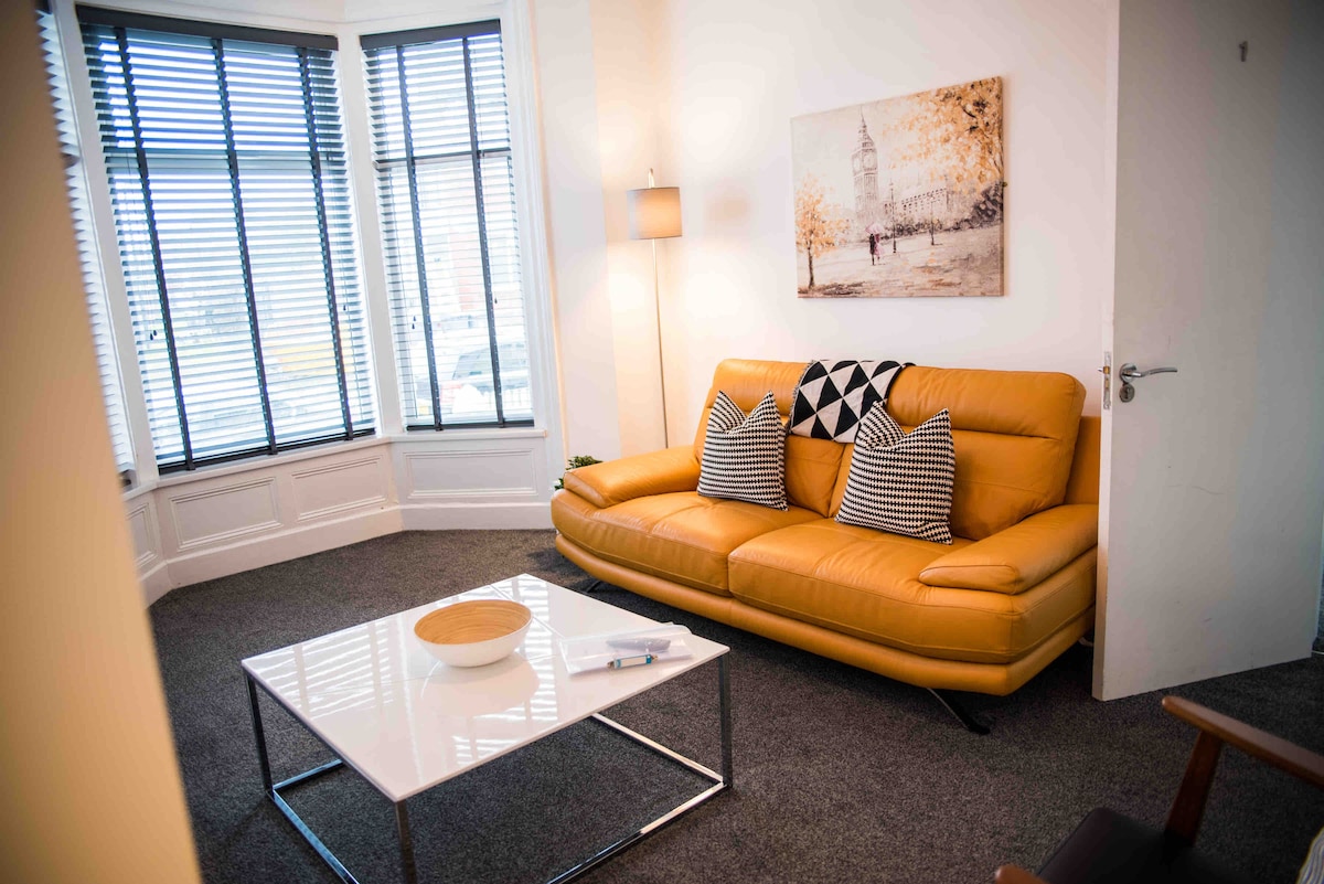 A bright living area is furnished with a modern yellow sofa, accented by patterned cushions. A sleek white coffee table sits in front. Large windows with blinds allow natural light to fill the space, while a wall art piece adds a touch of character.