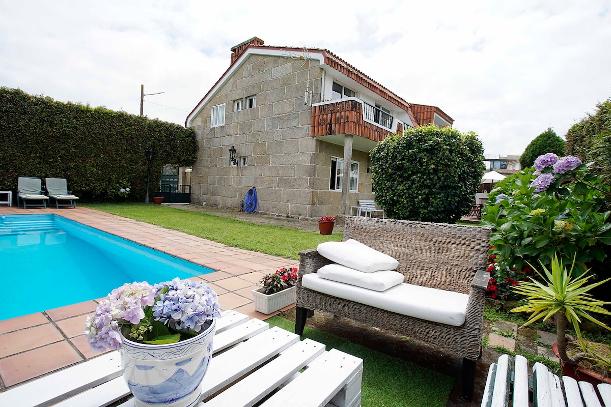 The outdoor area features a crystal-clear pool bordered by a manicured lawn, complemented by a white wicker seating arrangement. A stone house stands in the background, surrounded by lush green hedges and blooming flowers, creating a serene environment.