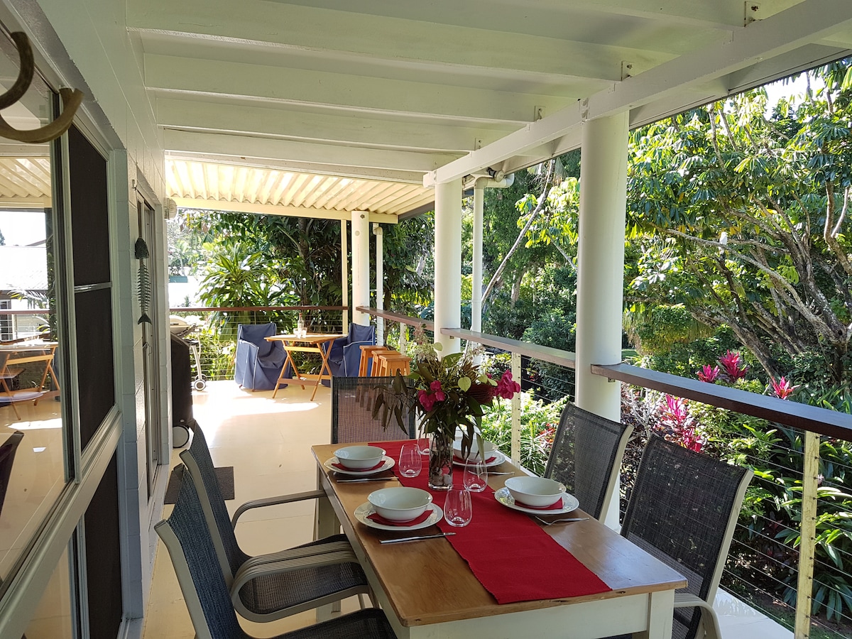 An outdoor dining area features a wooden table set for six, adorned with glassware and floral arrangements. Surrounding tropical greenery provides a lush backdrop, while seating options are available for relaxation nearby. The space is partially covered, allowing for comfortable dining in various weather.