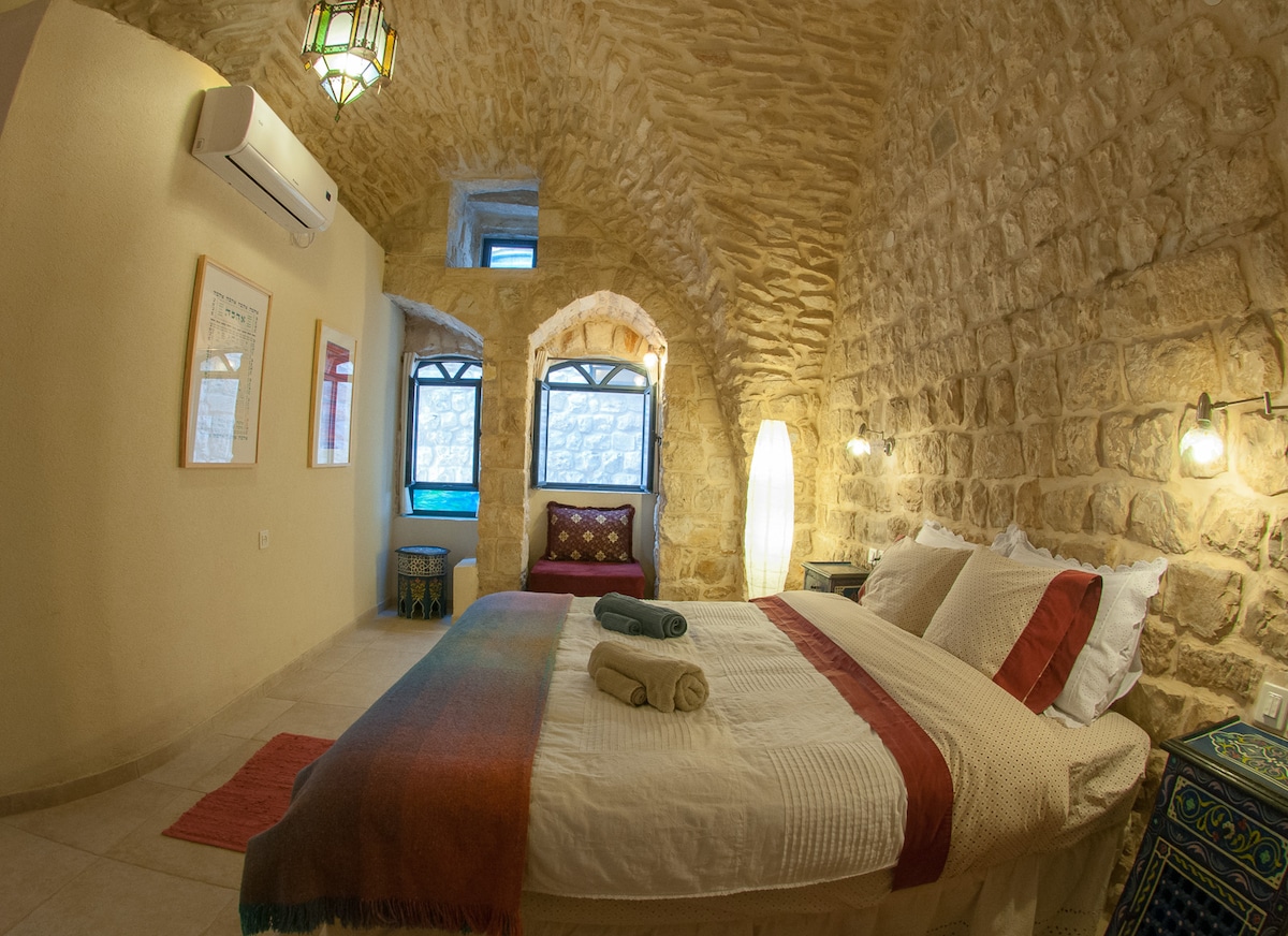 A stone-domed room showcases a spacious bed dressed in white with red and brown accents. Soft lighting is provided by wall sconces and a floor lamp. Windows with arches allow natural light, while a cozy seating area is visible in the background.