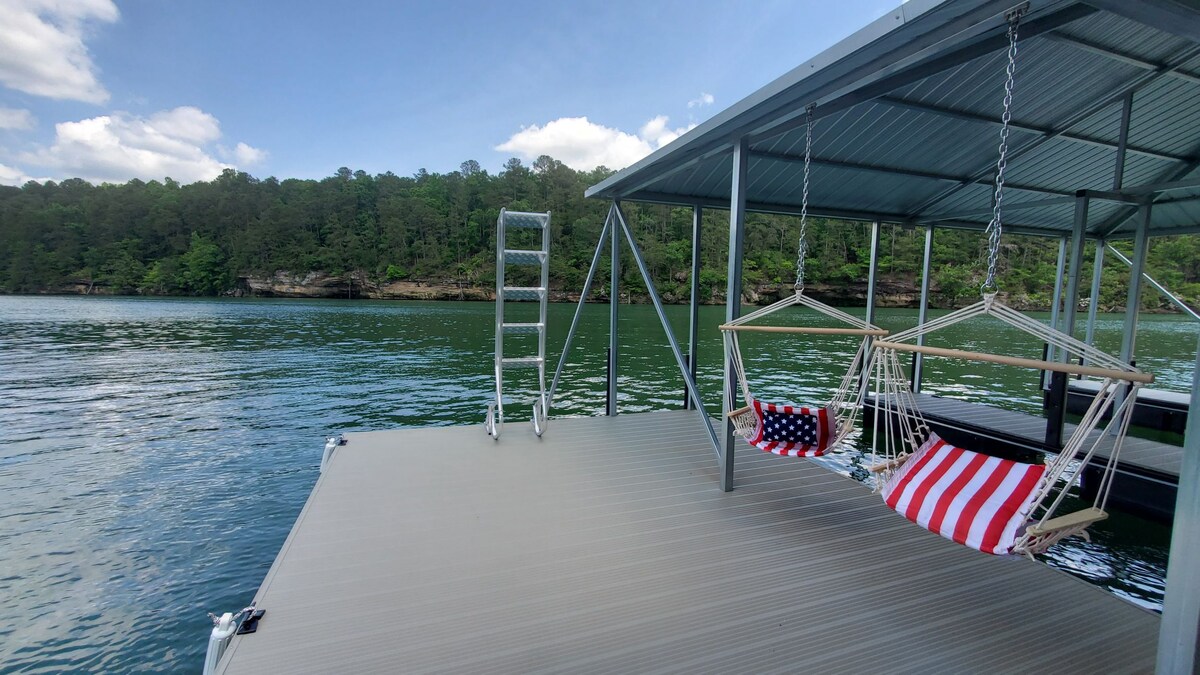 A covered two-slip dock is presented on calm waters, featuring swings adorned with patriotic stripes. A ladder is accessible for easy entry or exit, while lush greenery sets a serene backdrop. The surface is sturdy and allows for leisure and relaxation by the water.