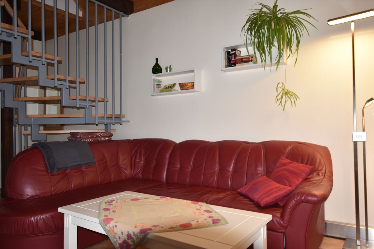 A cozy living area features a large red sectional sofa adorned with colorful throw pillows. A light-colored coffee table sits in front, while a plant hangs nearby. A staircase with wooden steps and a metal railing adds dimension to the space.