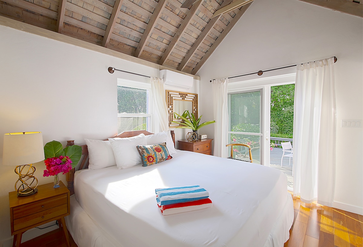 The bedroom features a spacious bed with crisp white linens and colorful accent pillows. Natural light pours in through large glass doors, leading to an outdoor deck. Two bedside tables hold lamps, with decorative plants adding a touch of greenery to the serene setting.