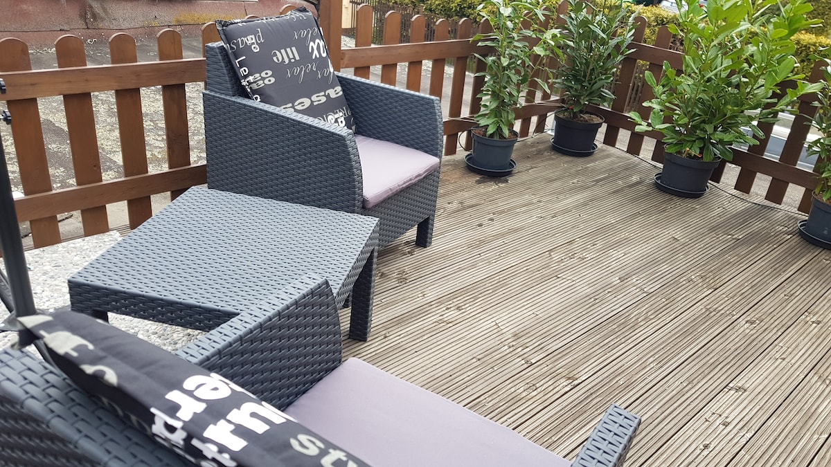 A wooden deck area features outdoor seating with dark wicker chairs and a matching side table. Potted greenery adds a touch of nature, providing a private space for relaxation. The surrounding wooden fence creates a cozy, enclosed atmosphere.