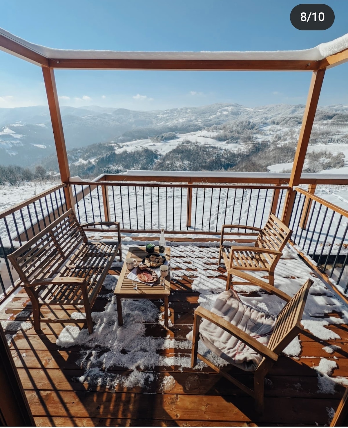 A wooden balcony is adorned with four outdoor chairs and a small table, partially covered in snow. The surrounding landscape features rolling hills and distant mountains under a clear blue sky, creating a serene setting for relaxation.