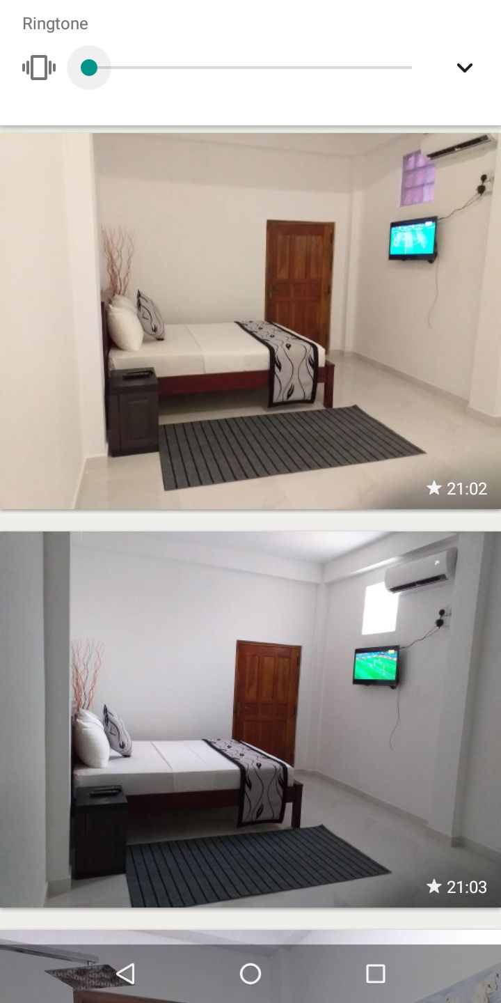 A bright bedroom features a spacious bed with a dark sheet and a decorative throw. A flat-screen TV is mounted on the wall. A window allows natural light to enter, and a simple area rug is positioned on the tiled floor.