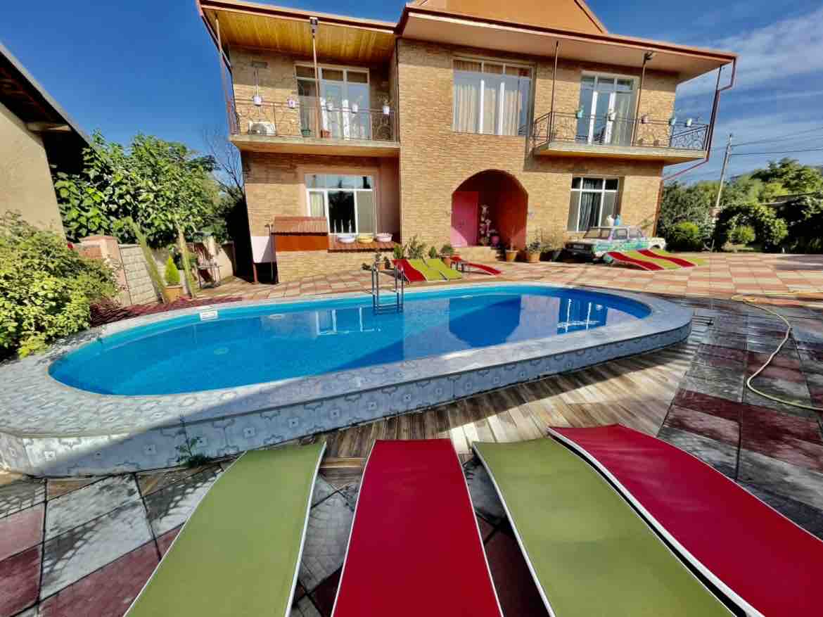 An outdoor swimming pool is surrounded by colorful lounge chairs in shades of red and green. The villa's two-story facade is visible in the background, with large windows inviting natural light. Lush greenery and garden plants frame the area, enhancing the serene atmosphere.