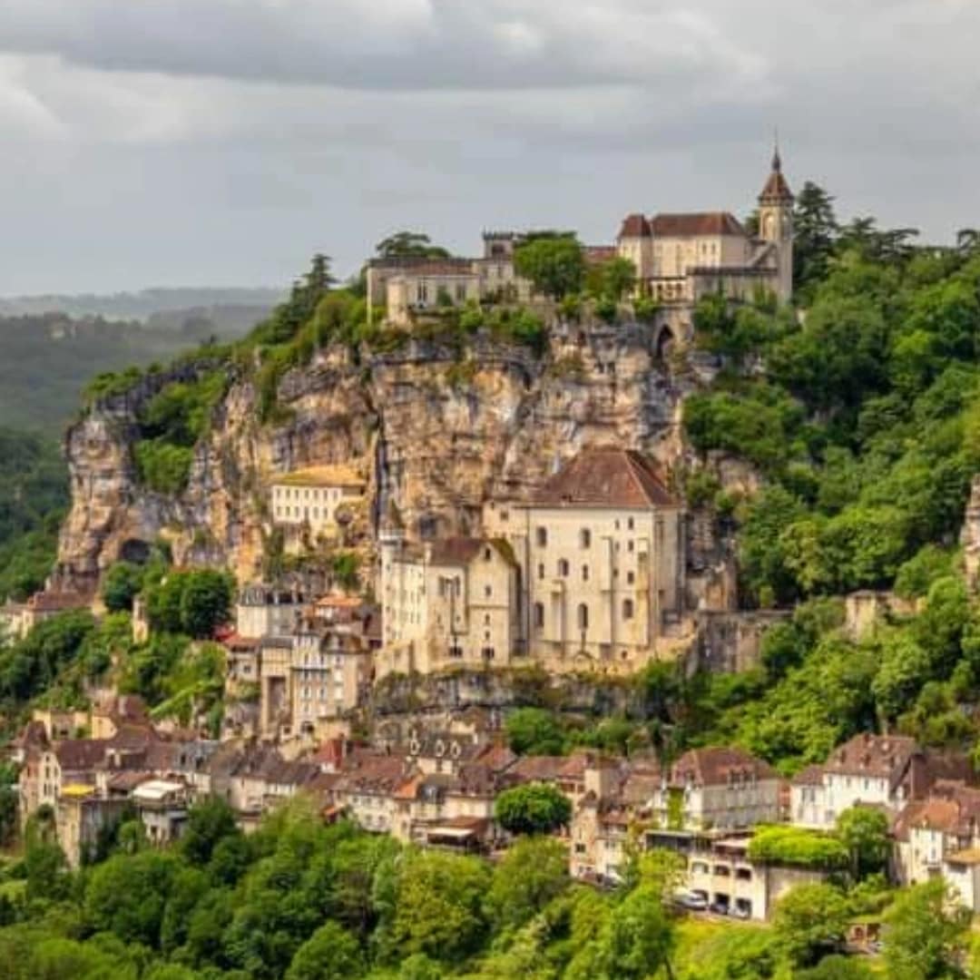 A scenic view of a rocky cliffside adorned with historic buildings and greenery. Several structures, including a church and a castle, are visible at varying heights, showcasing a rich architectural blend against the backdrop of rolling hills and cloudy skies.