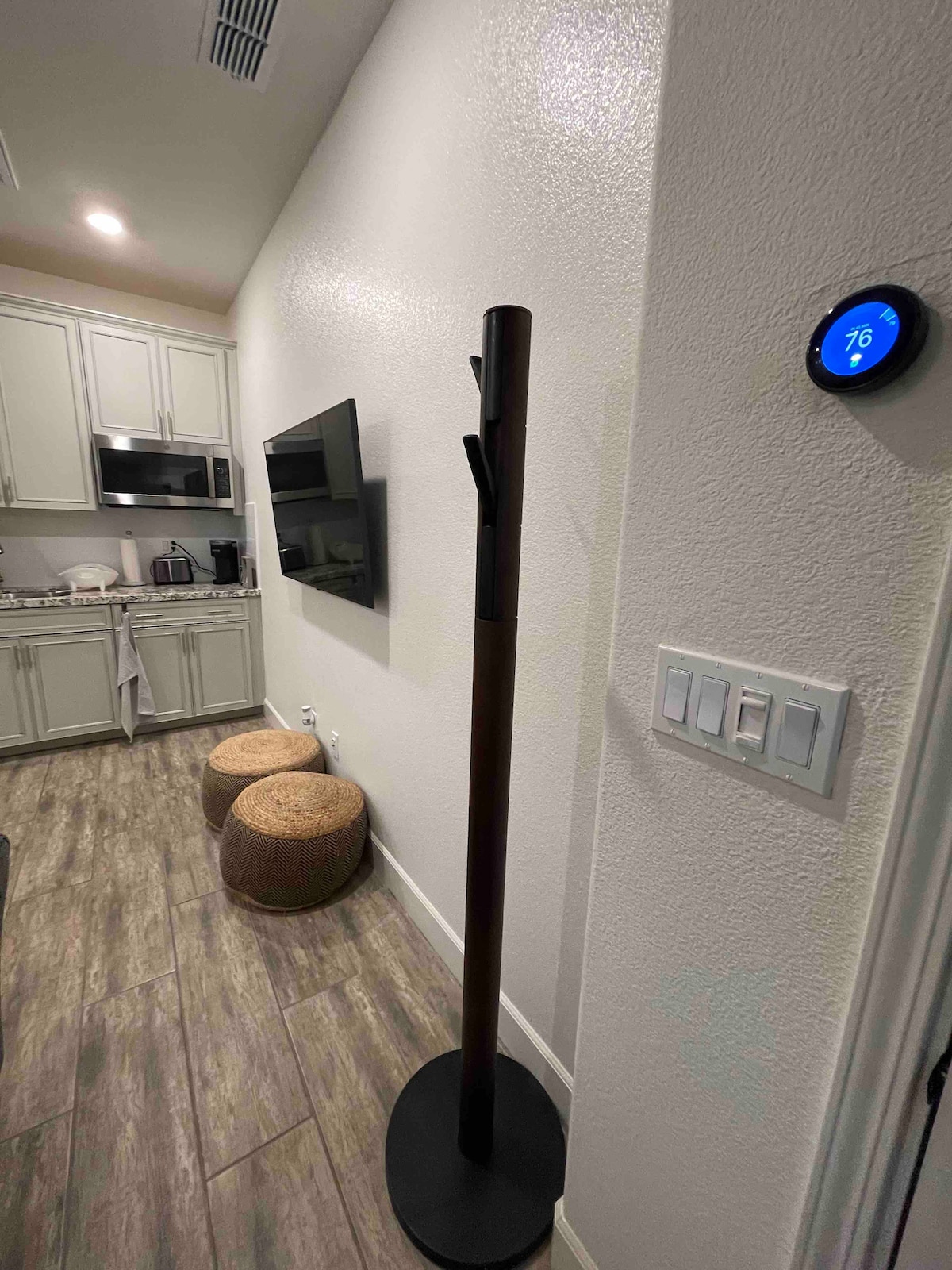 The image captures a corner of the studio featuring a tall, wooden coat rack positioned beside a modern thermostat. A flat-screen TV is mounted on the wall, facing a kitchenette outfitted with light cabinetry and appliances. Two round stools are placed in front of the kitchen area.