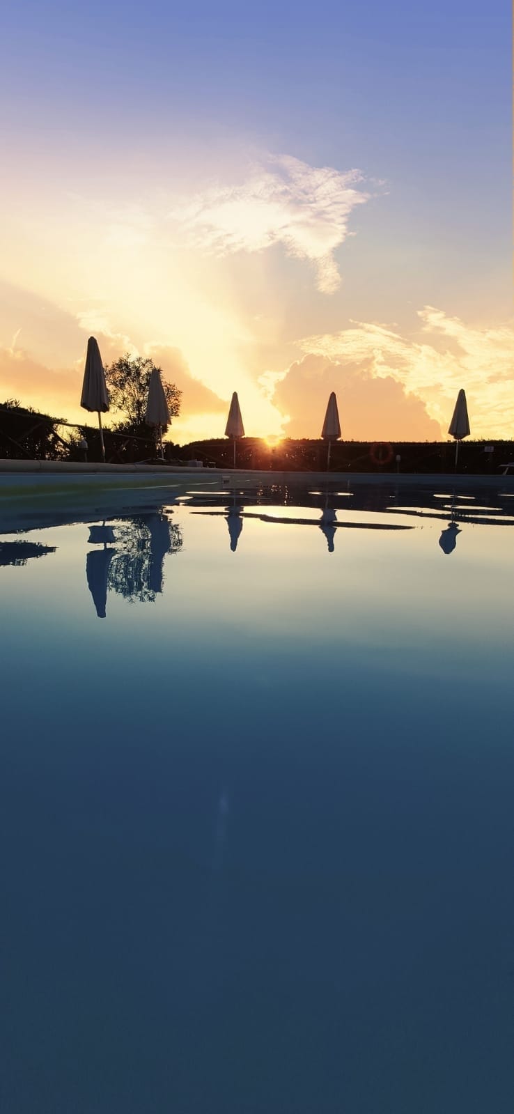 The image captures a serene pool at sunset, with gentle waves reflecting the vibrant colors of the sky. Umbrellas stand elegantly around the pool, enhancing the tranquil setting. A warm glow from the horizon complements the peaceful atmosphere of the villa.