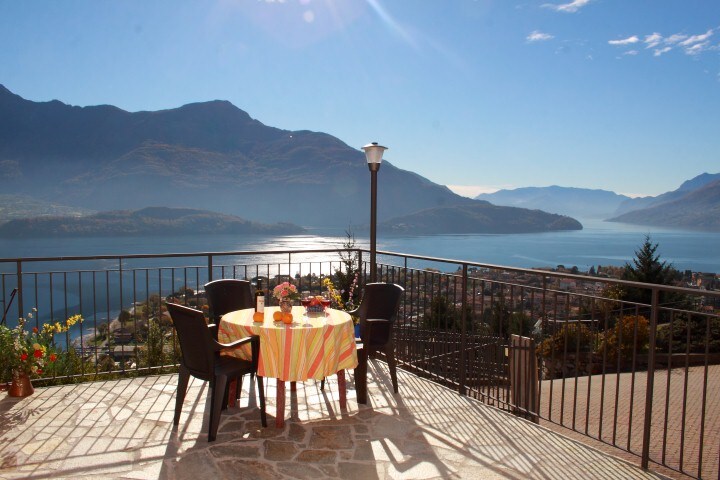 An outdoor terrace is presented with a round table set for two, adorned with a decorative tablecloth and flowers. Surrounding chairs offer additional seating. In the background, the calm waters of the lake reflect the mountains under a clear blue sky, enhancing the serene landscape.