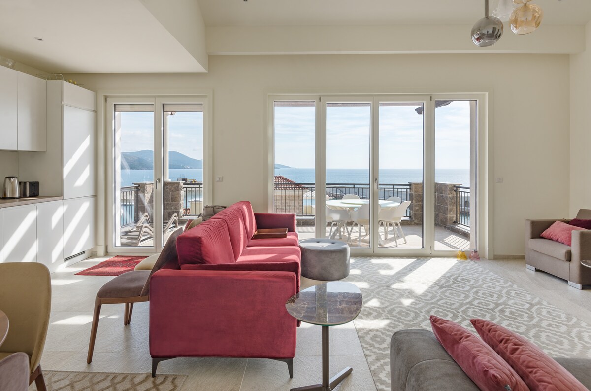 A bright and airy living space features a large seating area with a red sofa and a grey armchair. Large glass doors offer views of the sea and outdoor dining table. The area is illuminated by natural light and designed with neutral tones and patterned rugs.