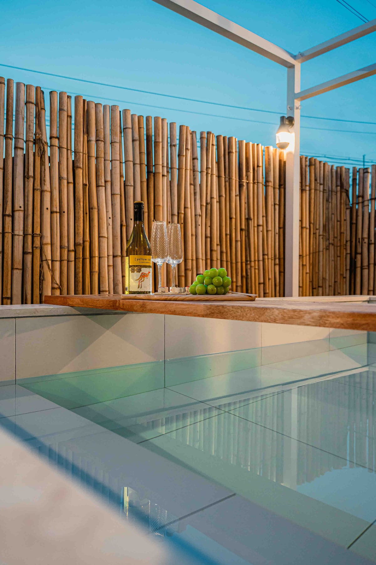 The outdoor jacuzzi is surrounded by bamboo fencing, providing a sense of privacy. A bottle of wine and two glasses are set on a wooden edge, accompanied by a small bowl of grapes. Soft evening light reflects off the water's surface.