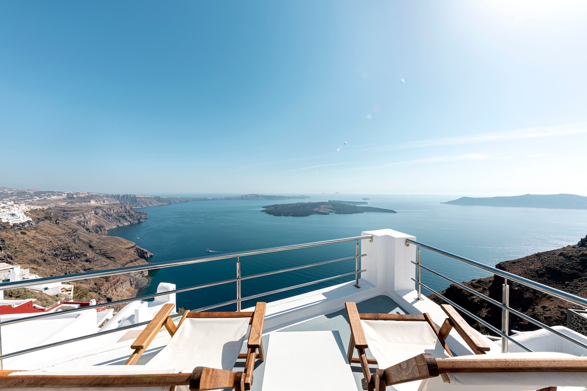 A large private terrace overlooks the stunning Aegean Sea, featuring two wooden lounge chairs positioned for relaxation. The clear blue sky blends effortlessly with the azure water, while the volcanic landscape is visible in the distance, creating a serene environment.