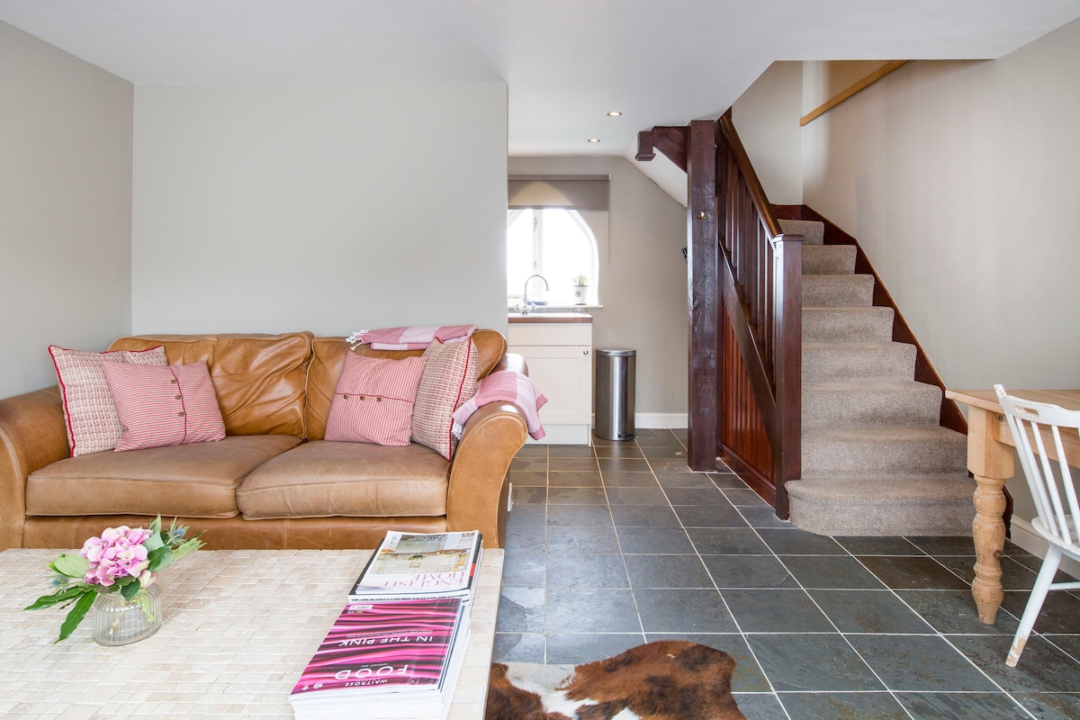 A cozy living area features a brown leather sofa adorned with pink cushions. A coffee table sits in front, displaying magazines and a small vase with flowers. A staircase leads to the upper level, while a glimpse of the kitchen is visible in the background.