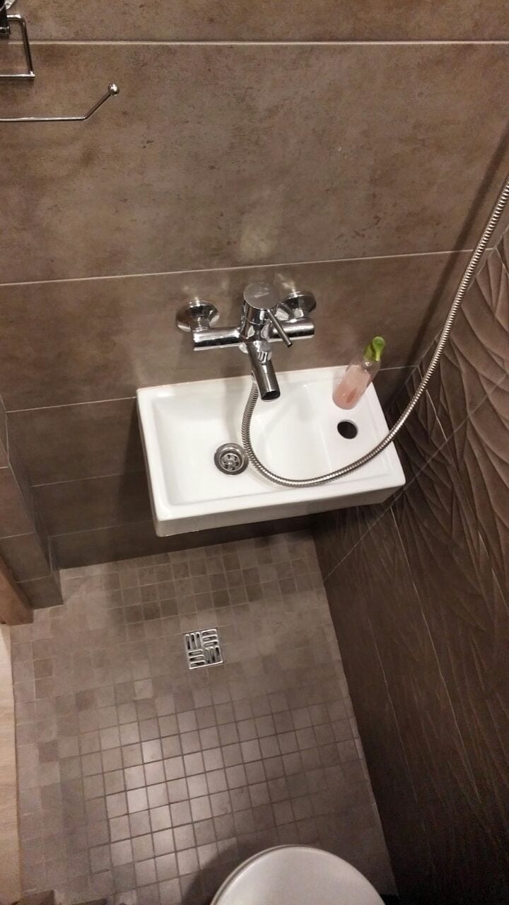 A compact bathroom space features a modern sink with a chrome faucet above a tiled floor. A handheld showerhead is attached to the wall, and a small bottle of toiletries is placed nearby. The walls are finished with neutral tiles for a clean look.
