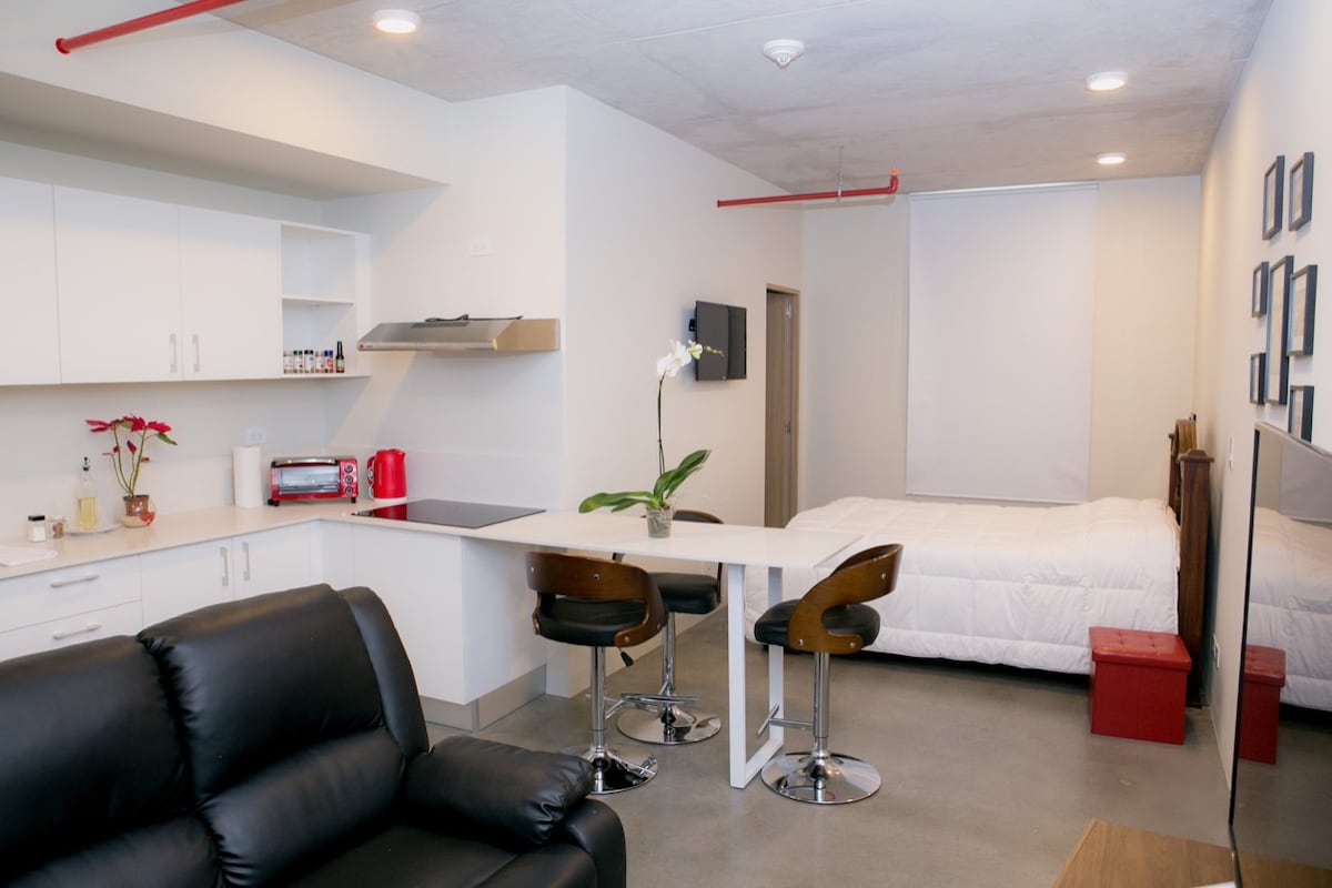 The open layout studio features a modern design with a spacious bed positioned alongside a kitchenette. A sleek counter with three bar stools provides seating. A comfortable black sofa faces a wall-mounted TV, while light colors enhance the overall brightness of the space.