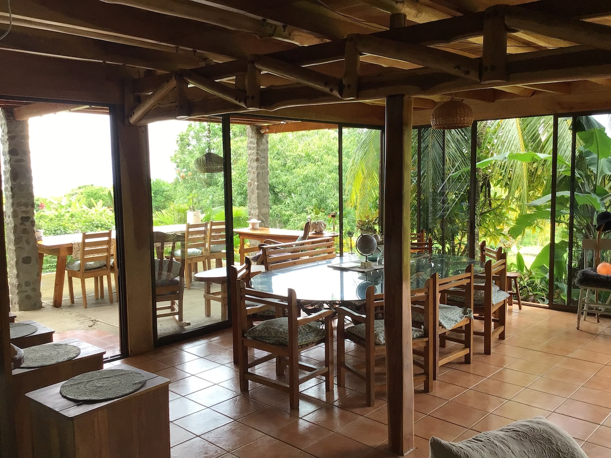 An open dining area features a large glass table surrounded by wooden chairs, with views through floor-to-ceiling glass doors. The natural light fills the space, complemented by surrounding greenery visible from the outdoor seating area.