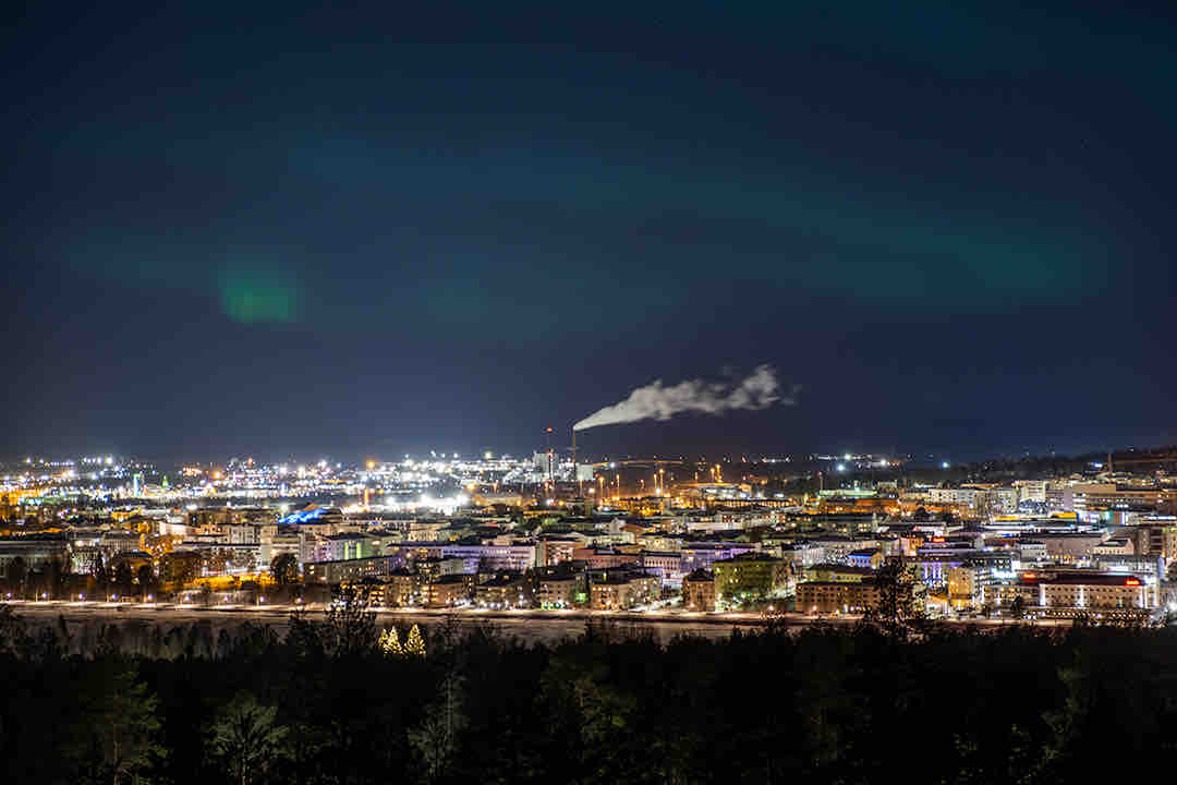 A night view of Rovaniemi showcases a vibrant city illuminated by numerous lights. Smoke rises in the distance, while the northern lights add a touch of color to the sky. The landscape reveals a mix of urban and natural elements, inviting exploration of the city.
