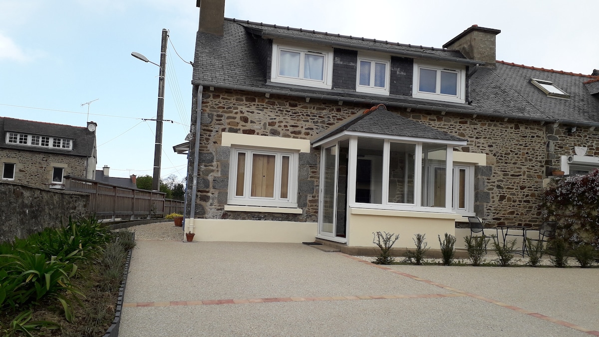 A charming stone house features a light-colored façade and a welcoming glass-enclosed entryway. The spacious driveway is bordered by neatly arranged plants, enhancing the exterior layout. Windows on the upper floor allow natural light to illuminate the interior.