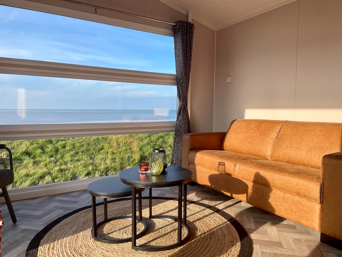 A cozy seating area features a brown leather sofa positioned next to two circular coffee tables. Curtains frame a large window offering a view of the Wadden Sea and surrounding greenery. Soft natural light brightens the space, creating a relaxing ambiance.