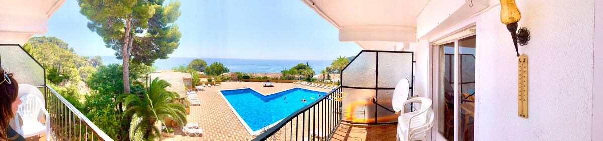 A panoramic view features a private pool surrounded by sun loungers, overlooking a serene sea. The balcony is furnished with a chair and a table, allowing for leisurely outdoor enjoyment. Lush greenery frames the scene, contributing to the tranquil atmosphere.