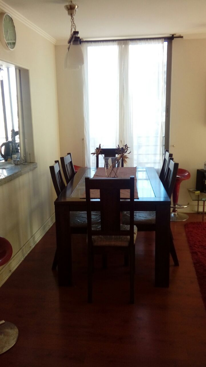 A dining area is equipped with a large wooden table surrounded by eight chairs. Natural light enters through two tall windows, which are adorned with sheer white curtains. A decorative centerpiece is placed on the table, complementing the warm wooden tones of the space.