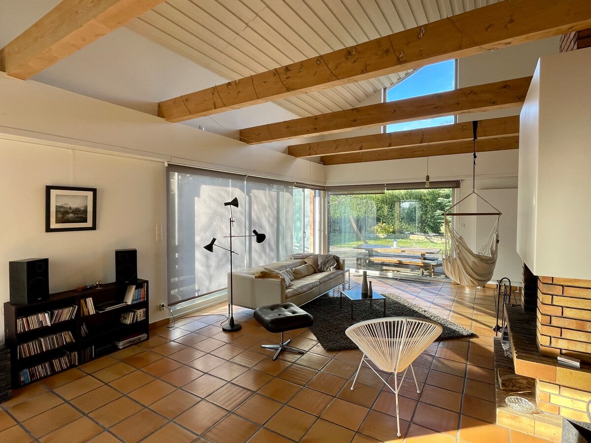 An airy living space features high wooden beams and large windows allowing natural light to fill the room. A comfortable lounge area includes a modern sofa and a stylish hanging chair. An inviting fireplace is situated next to a low table, offering a cozy gathering spot.